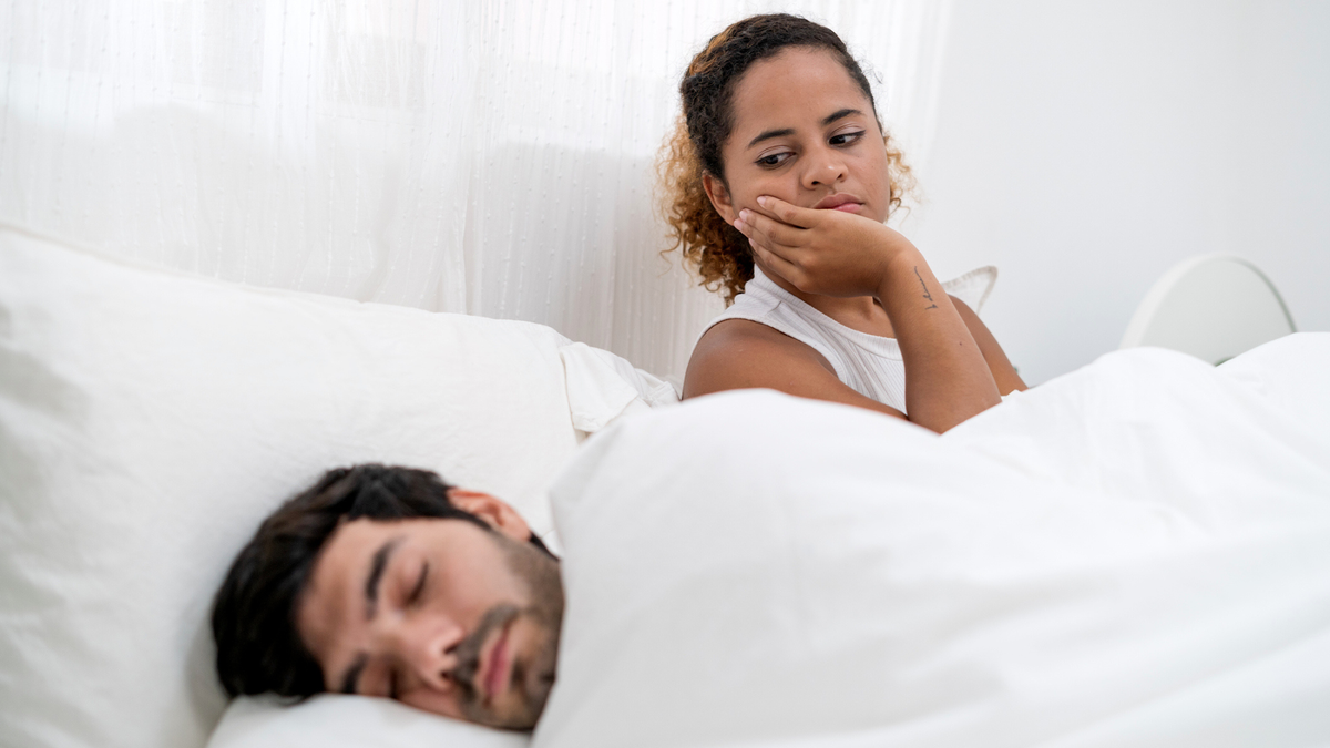 Man asleep next to partner who is sat up awake and looking at him with an annoyed expression because he&#039;s been talking in sleep, which keeps waking her up