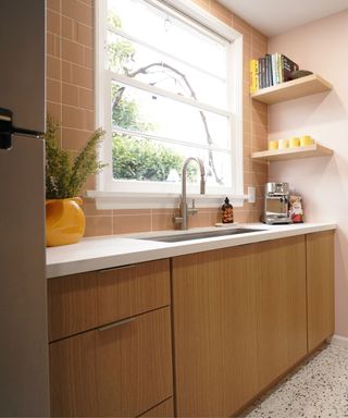 A kitchen with pale pink walls, a pale pink tile backsplash, wooden cabinets, and a small window.