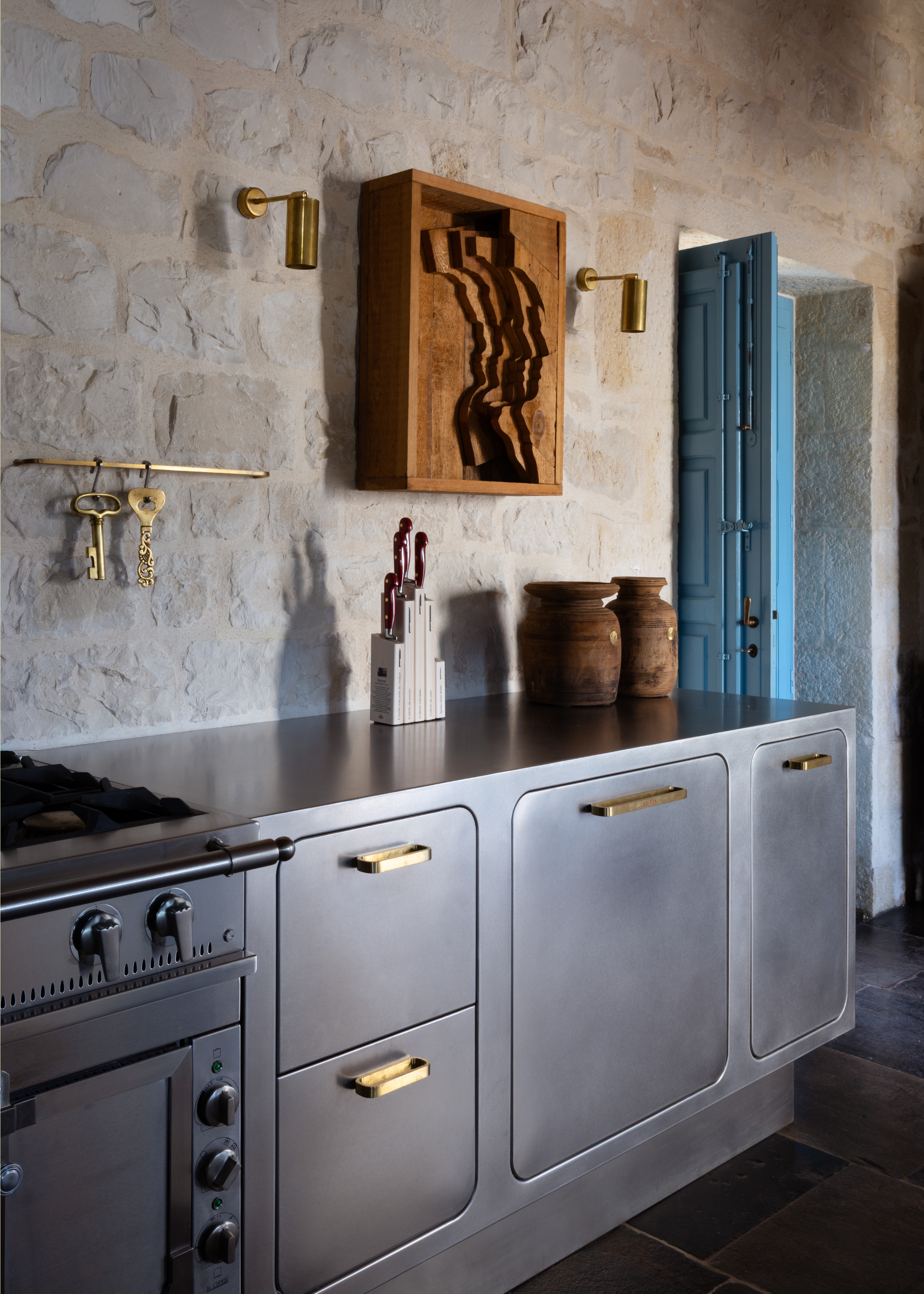 a kitchen with sanded steel cabinetry with gold hardware, stone walls, and wooden flooring