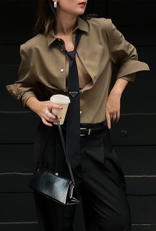 The frame bag trend is shown in a street-style photo of a woman wearing silver earrings, a brown button-down shirt, a black Prada tie, a black belt, black trousers, and a black frame bag