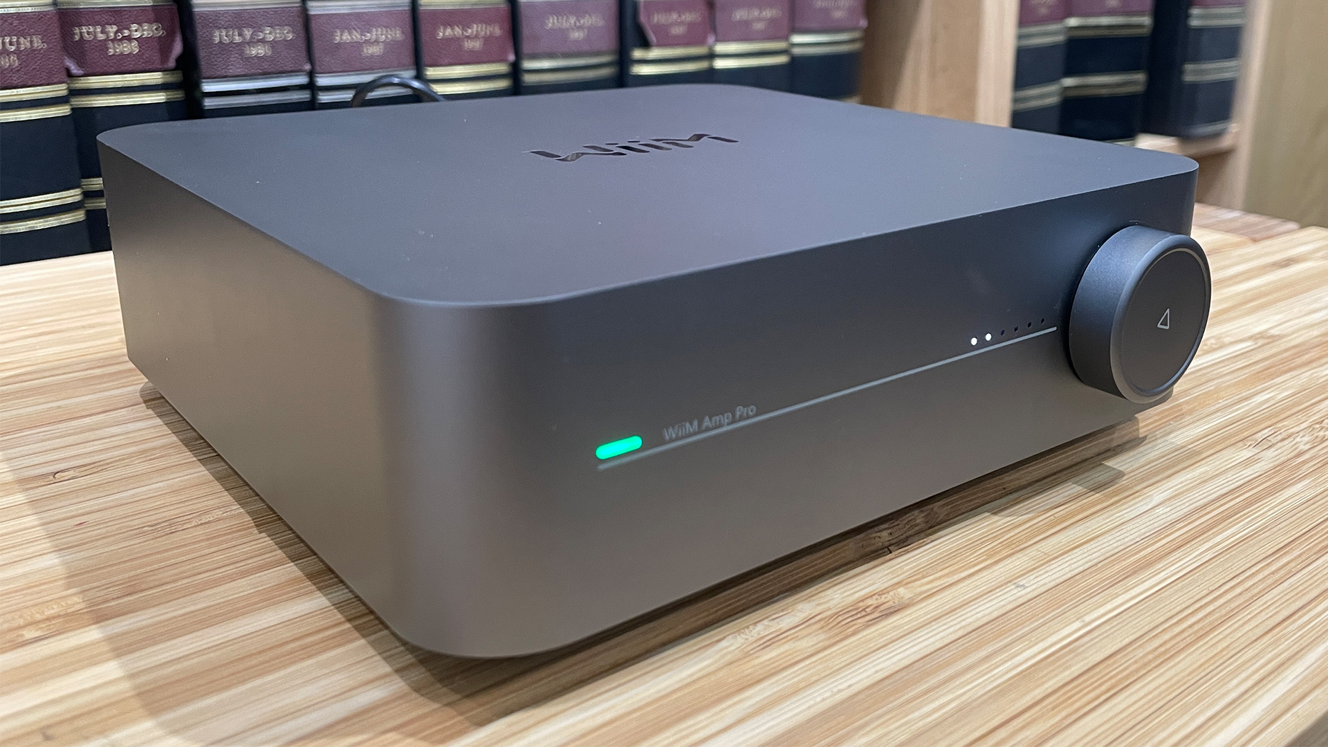 WiiM Amp Pro streaming amplifier on wooden hi-fi rack in front of bookcase slight angle view