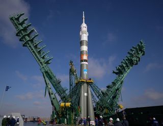 A green/grey rocket with a pointy white top stands surrounded by diagonal support arms, leaning as they are raised to encapsulate the launch vehicle for access before launch.