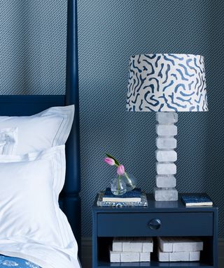 blue and white bedroom with blue four posted bed and bedside table, patterned wallpaper and patterned shade