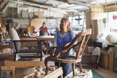 A portrait of an upholsterer in her workshop. She leans against a chair frame with her colleague working in the background.