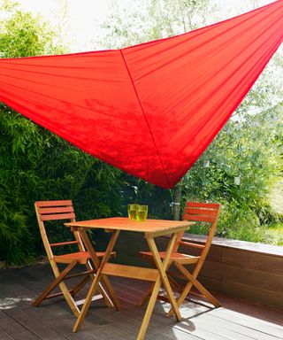 Red garden sail providing shade for wooden table and chairs on decked patio in garden