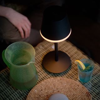 Illuminated black smart lamp on a rattan garden table with green glass pitcher and drink beside it