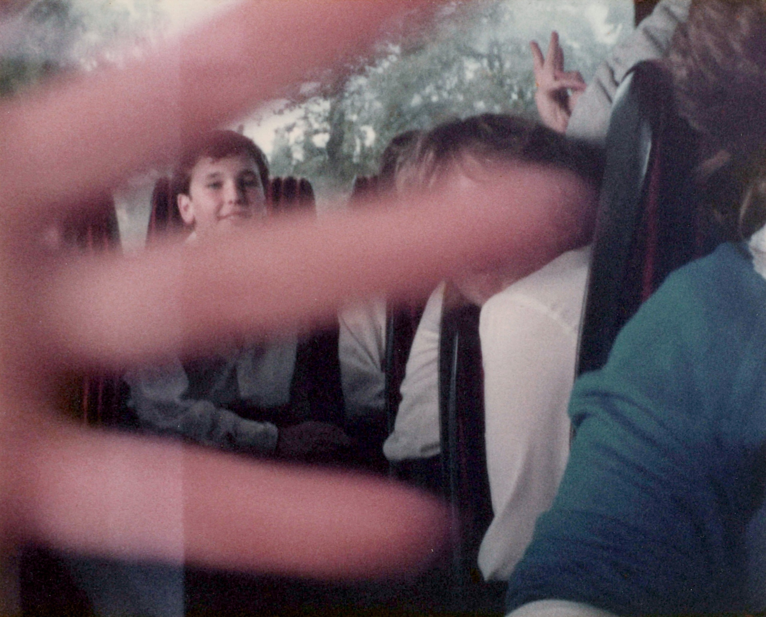 A hand in front of the lens obscures a scene of children on a coach, with one child's face visible through the gaps in the fingertips