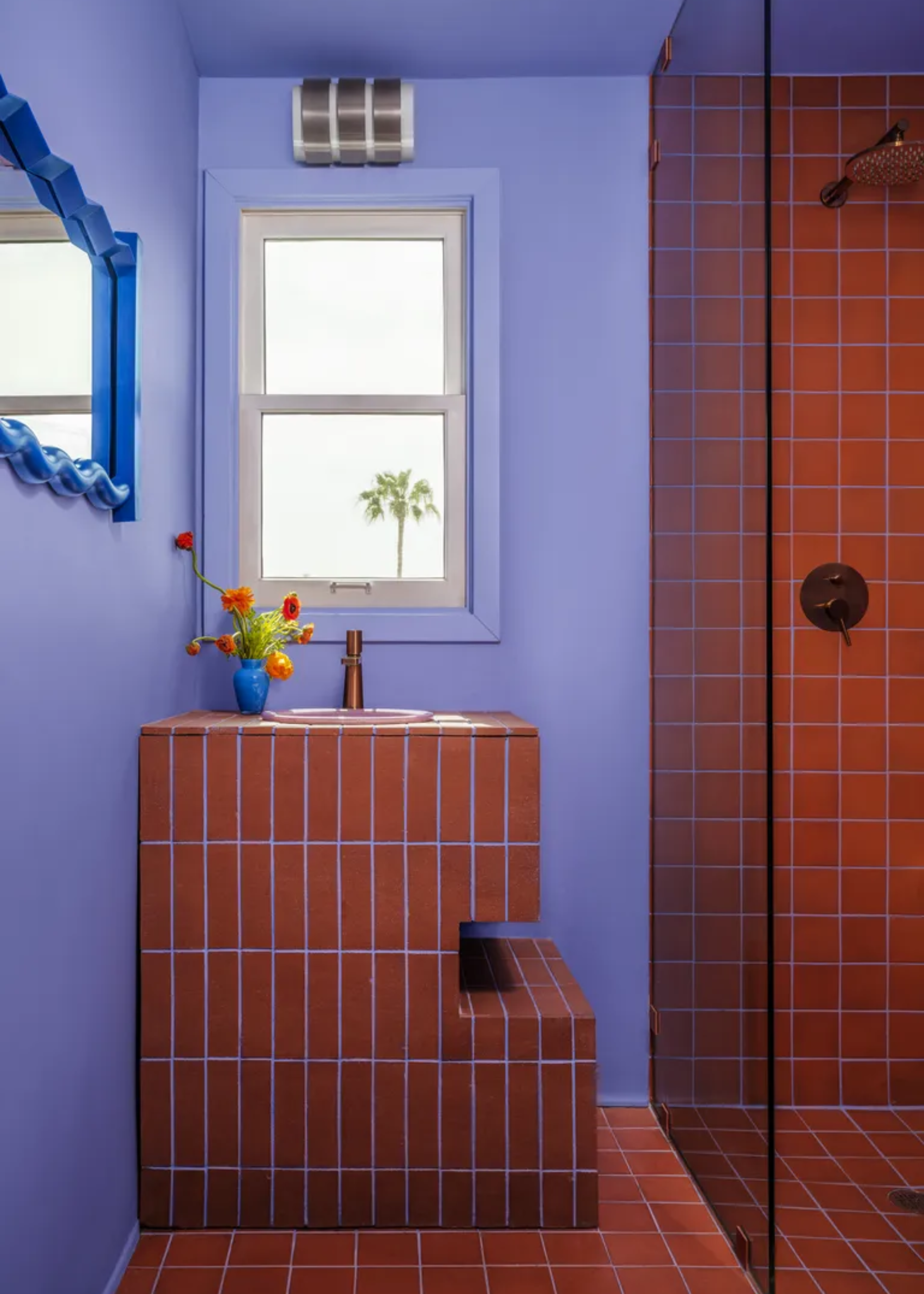 A small purple bathroom with terracitta-colored tiles and purple grout by a small window, wavy blue mirror and a shower room