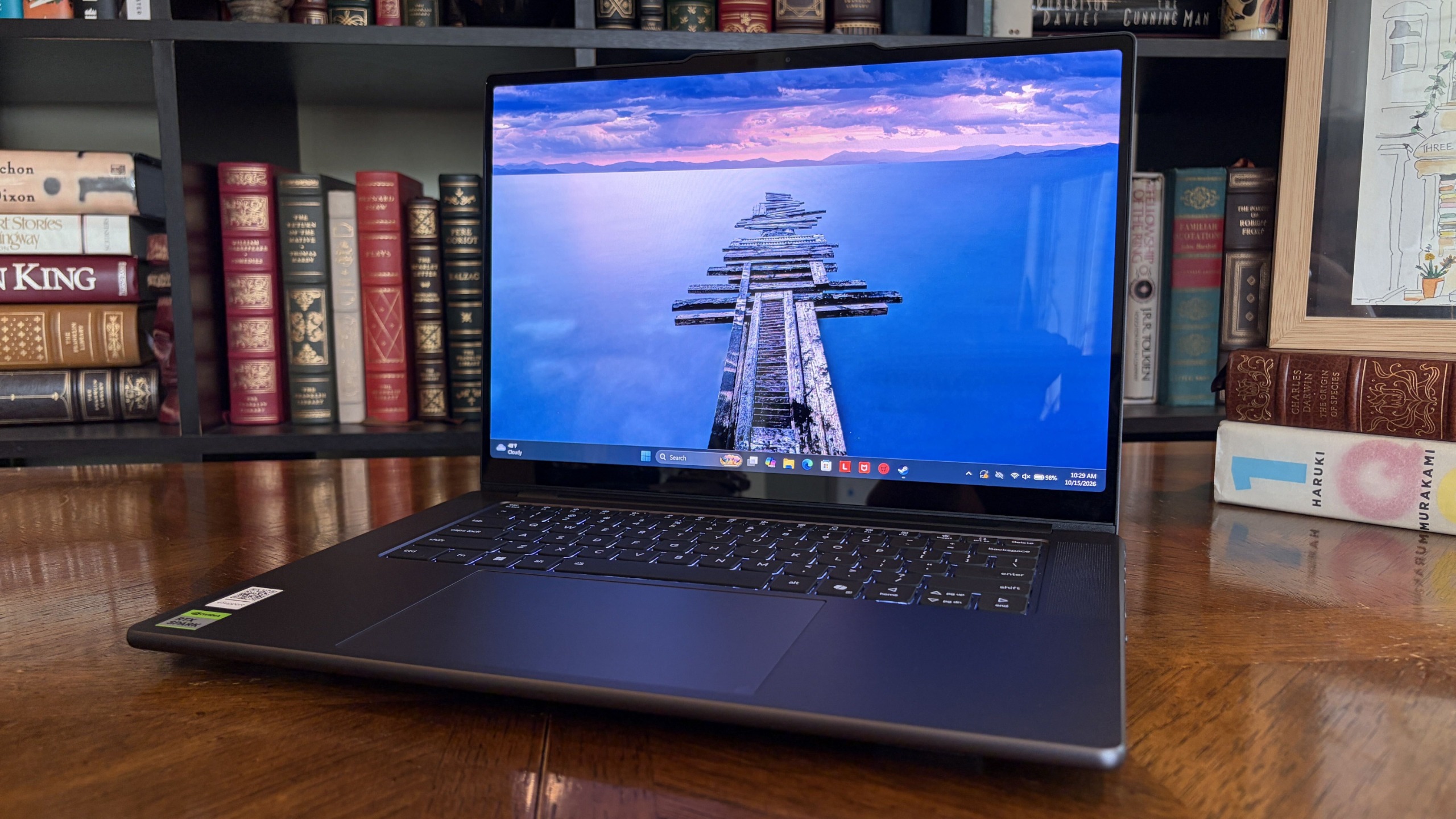 A slim Lenovo Pro 9n laptop rests on a polished wooden desk before bookshelves, its screen showing a serene blue ocean and pier beneath a pink sunset.