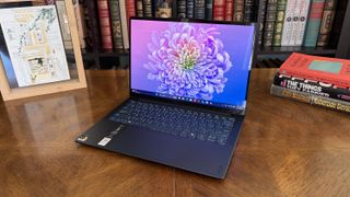 A sleek laptop with a vibrant purple flower on the screen sits on a wooden table. Books and framed art surround it, evoking a cozy, intellectual atmosphere.