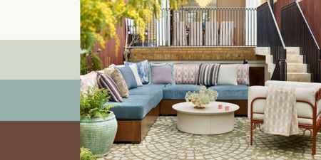 An outdoor living space with a cornflower blue corner sofa, lilac patterned cushions, a cream round coffee table, and a modern minimalist armchair with a rust-colored frame and cream cushion. 