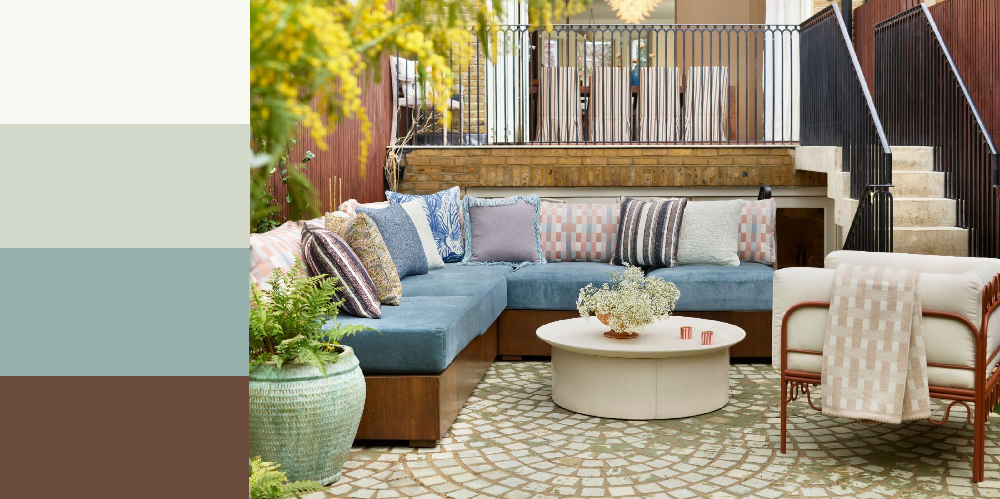 An outdoor living space with a cornflower blue corner sofa, lilac patterned cushions, a cream round coffee table, and a modern minimalist armchair with a rust-colored frame and cream cushion. 