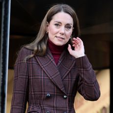 Kate Middleton wearing a plaid coat and red turtleneck lifting one hand up to her face