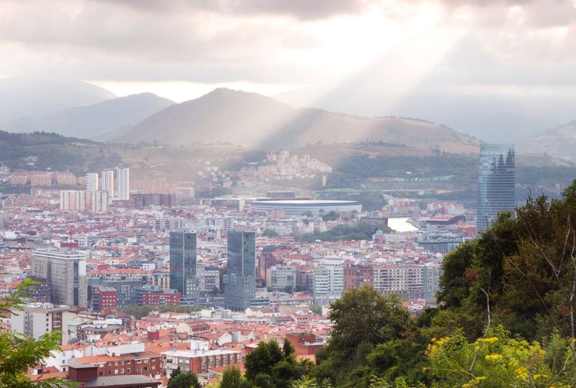A panoramic view of the Basque Country showcasing a cityscape with mountains in the background, illuminated by rays of sunlight.