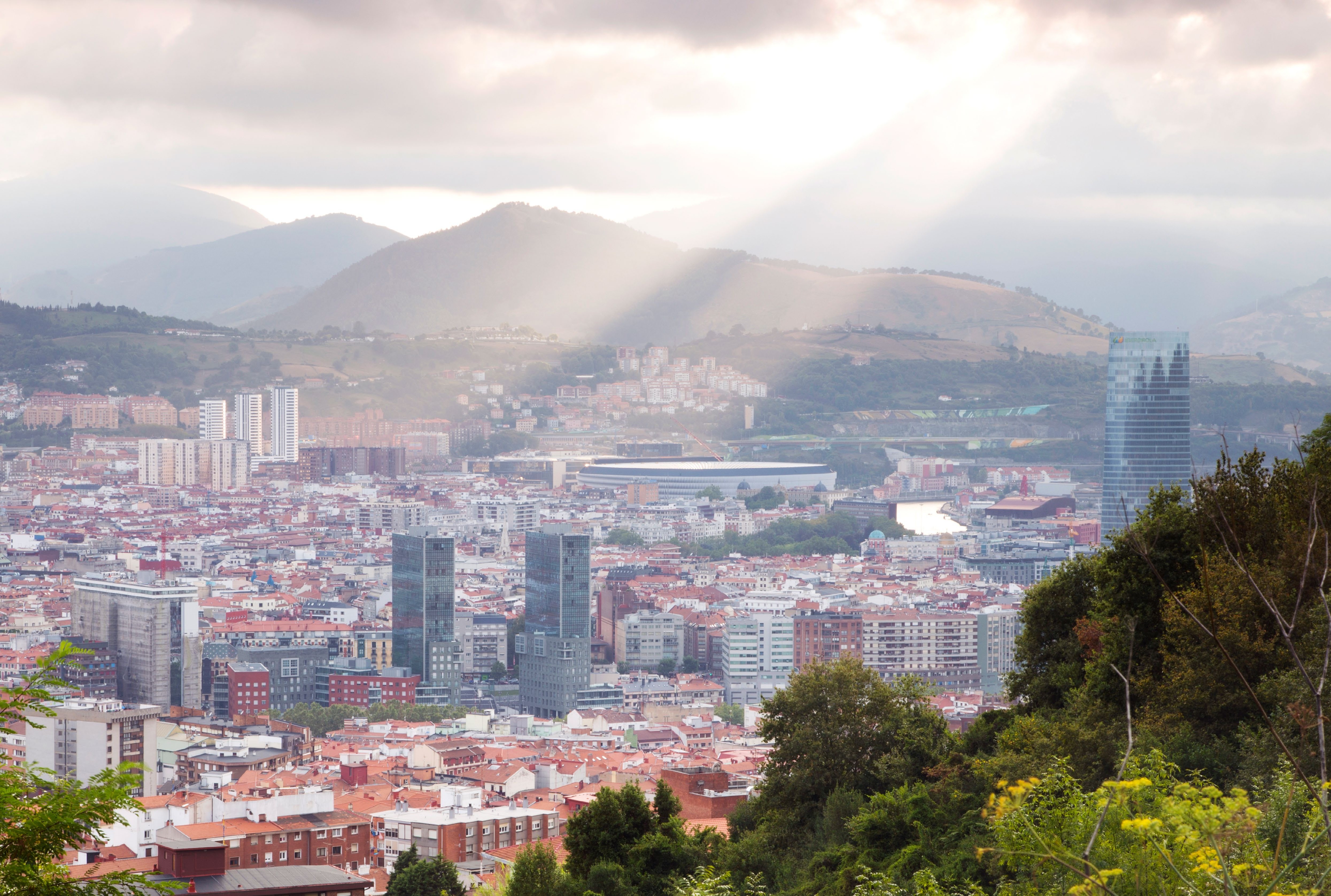 A panoramic view of the Basque Country showcasing a cityscape with mountains in the background, illuminated by rays of sunlight.