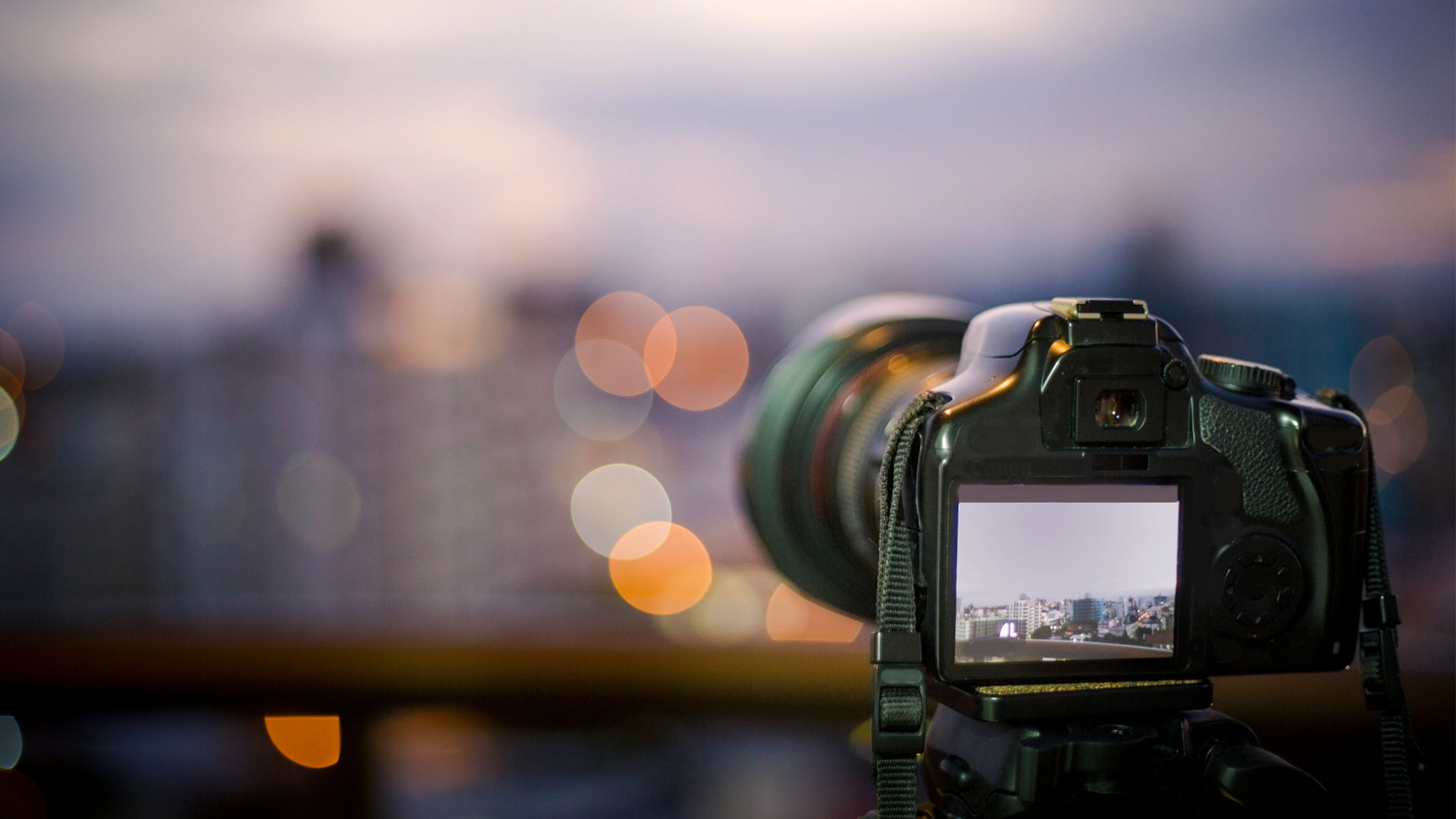 DSLR fotoaparatas, pastatytas prieš saulėlydį su vaizdu į nefokusuotą miestą
