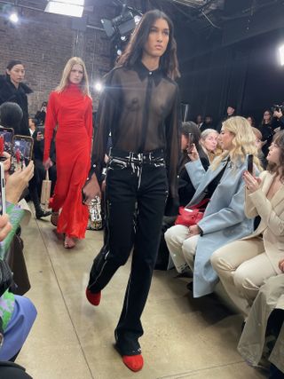 Proenza Schouler model wearing a sheer black top, black jeans, and red flats