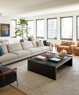 A living room with a large beige sectional, vintage armchairs and a tall tree in the corner