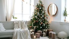 A Christmas tree with presents in a small living room with a gold mirror and a white couch. 