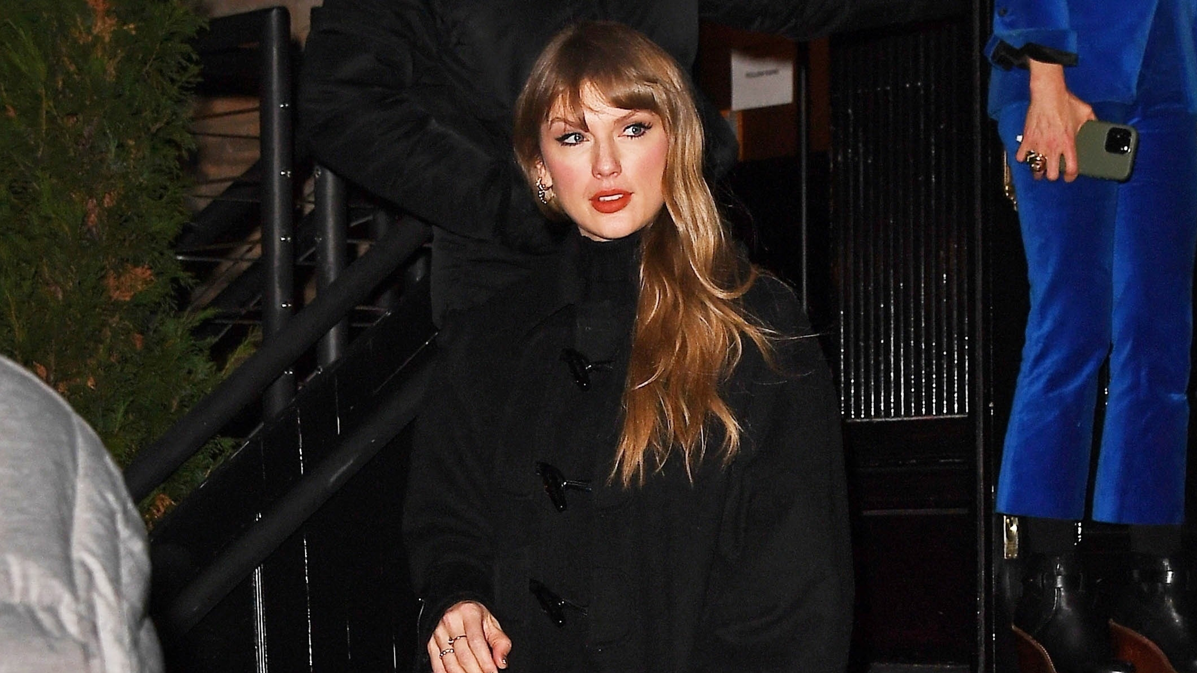 Taylor swift wears a black wool coat, black sheer tights, and black knee-high boots.