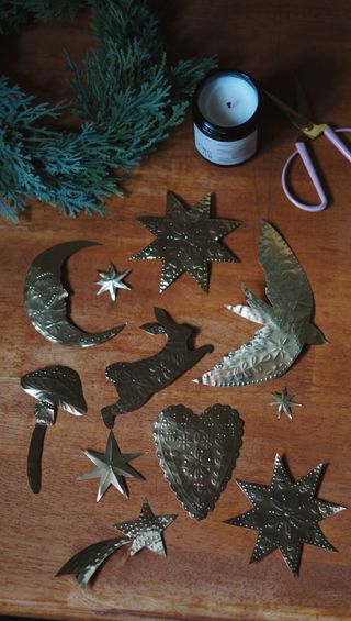 Several Gold Embossed christmas tree ornaments on a wooden table with a wreath in the top left corner and a candle