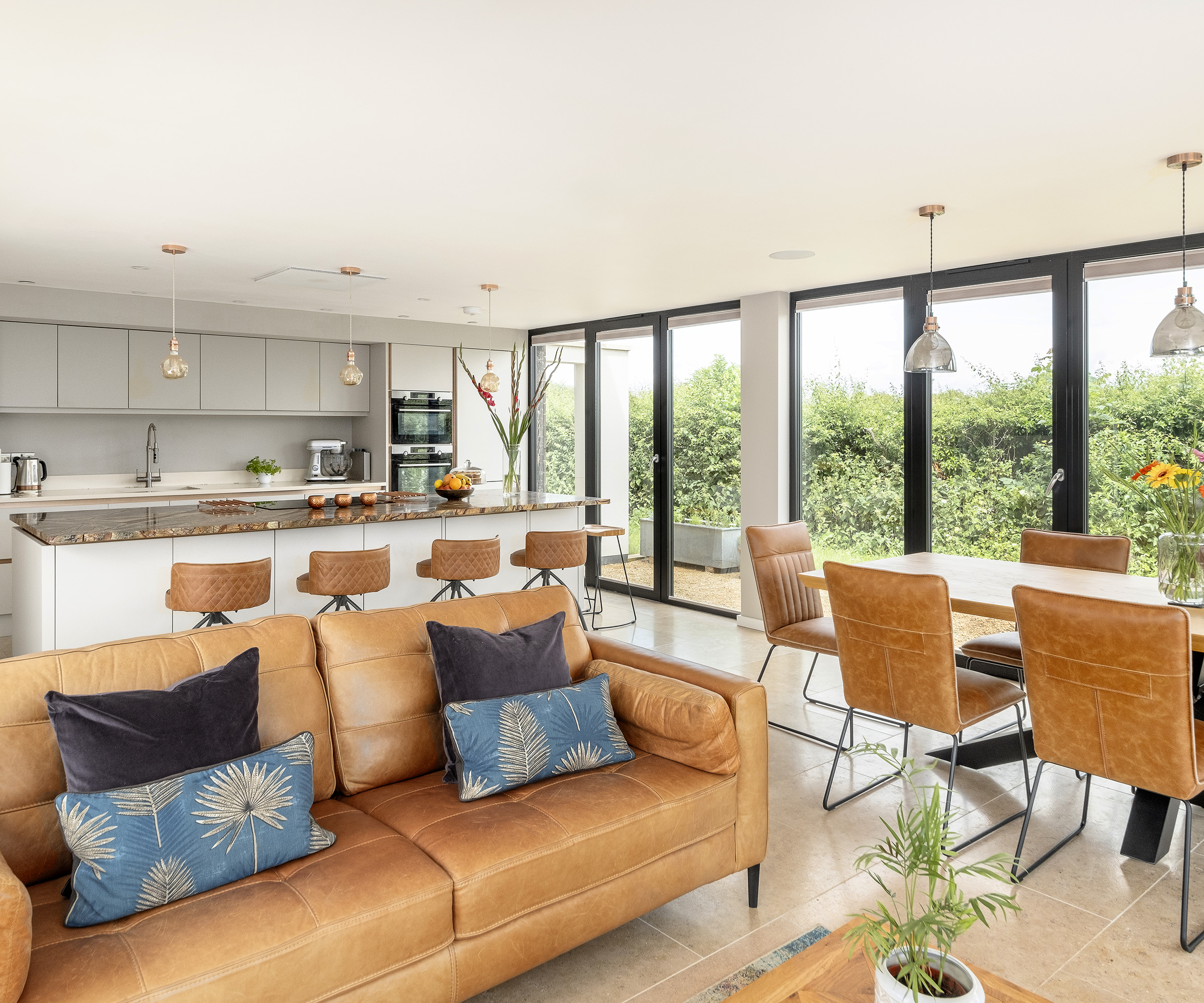 A multi functional kitchen-diner-lounge space with tan coloured furniture and a wall of floor to ceiling glazed doors