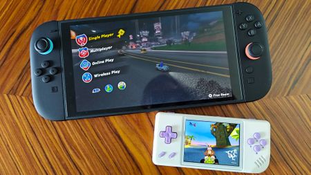 Anbernic RG28XX handheld with Crash Team Racing on screen sitting under Nintendo Switch 2 running Mario Kart World on woodgrain table.