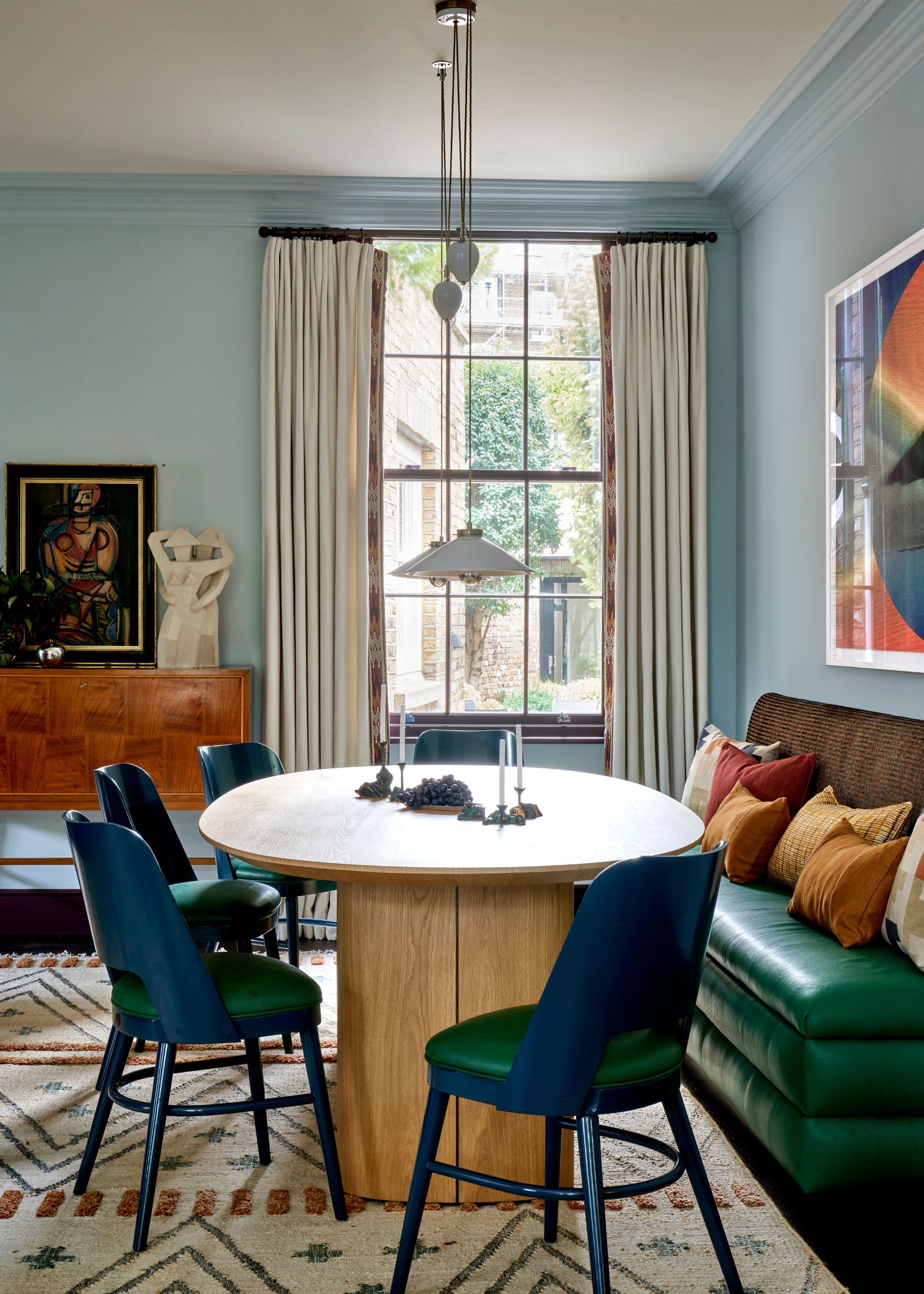A living-dining room with pale blue walls, green bench seating, and a modern wooden dining table with blue and green chairs. A patterned cream rug, cream curtains on the window, and abstract artwork on the wall.