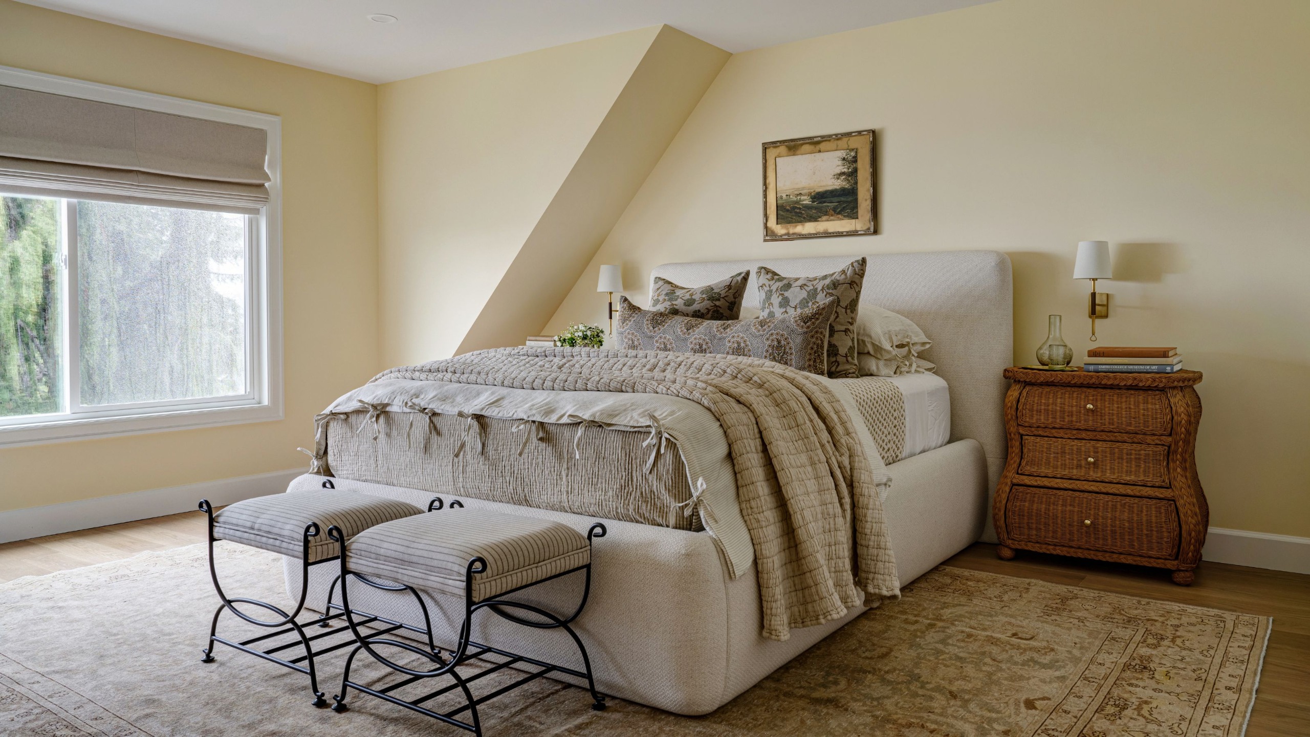 A calming bedroom with pale yellow walls, a vintage rug, a neutral fabric headboard, and lots of neutral layered bedding. Two small upholstered seats at the end of the bed, a wooden nightstand, and vintage artwork.