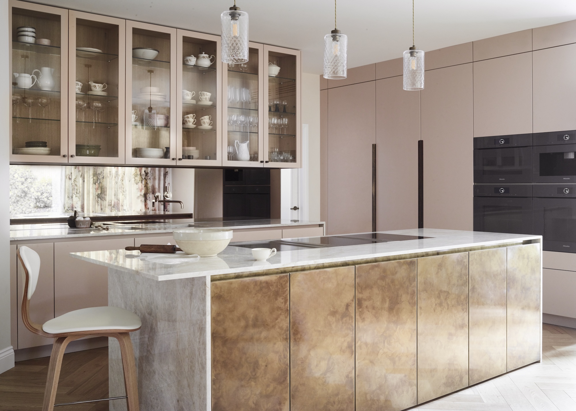 A kitchen with glass fronted cabinets and a large island in the middle with golden/metal fronts as well as three pendants hanging above it.