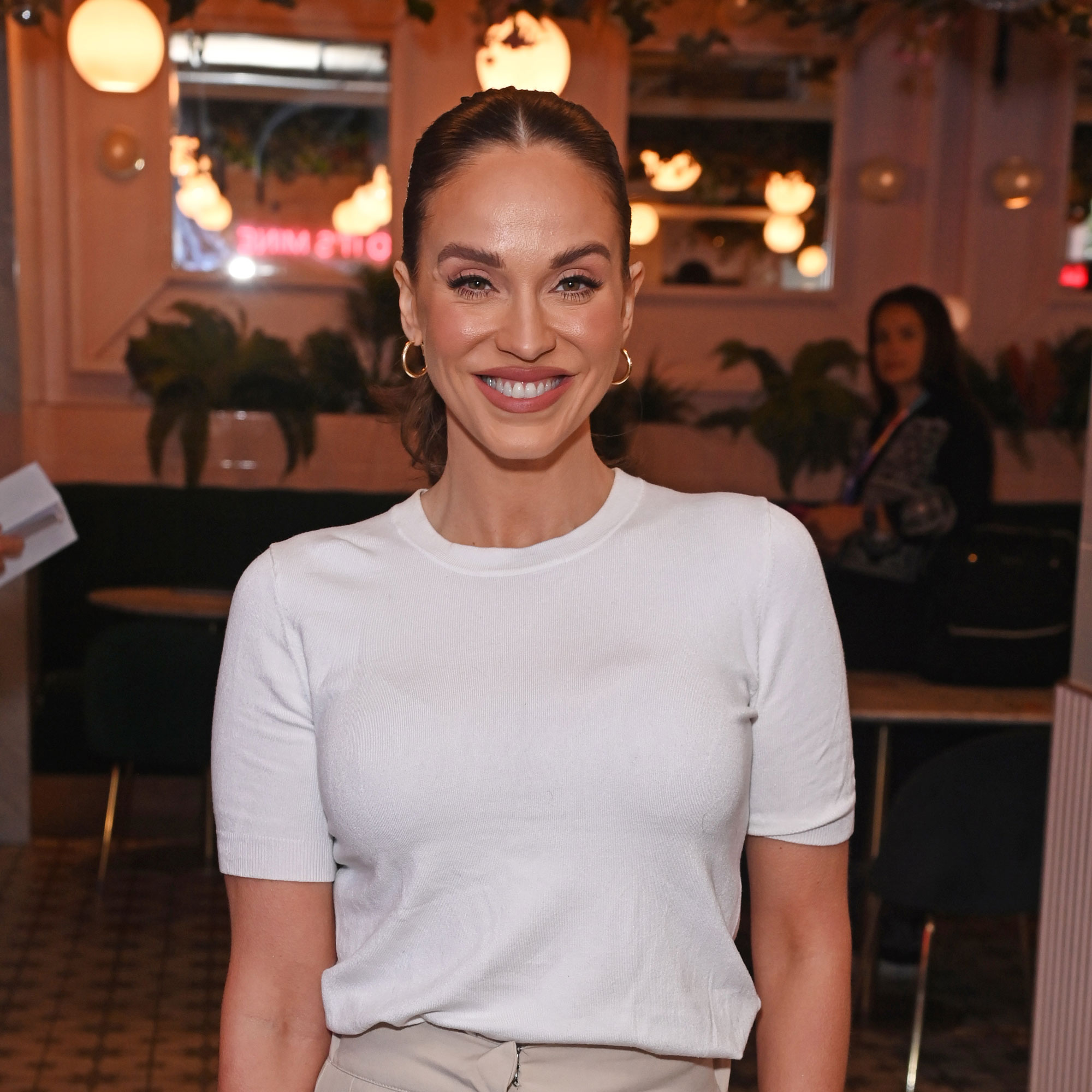 Vicky pattison wearing a white t-shirt, brown trousers, hair in a pony tail and gold earrings