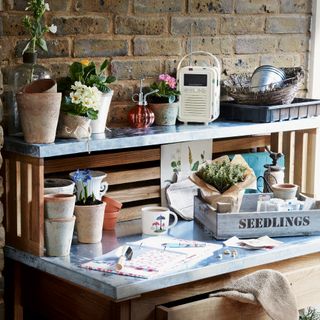 Metal potting bench against a brick wall, with stacked terracotta pots and a blue wooden tray labelled 'seedlings'