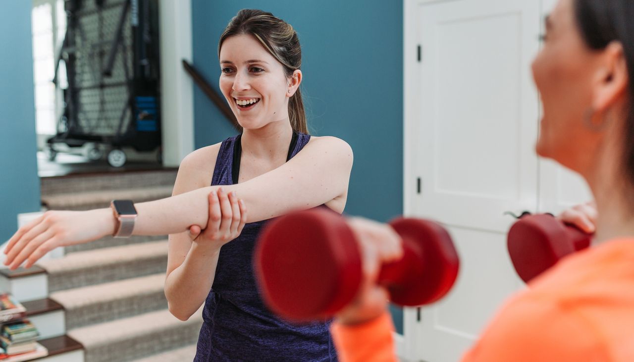 A smiling woman stretches her arm across her chest. A women in an orange top in the foreground holds red dumbbells by her shoulders