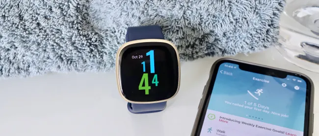 Un smartwatch Fitbit Versa colocado sobre una superficie blanca junto a un teléfono móvil que muestra una aplicación de ejercicio en la pantalla. Ambos dispositivos están junto a una toalla gris, destacando el ambiente de fitness y actividad física.