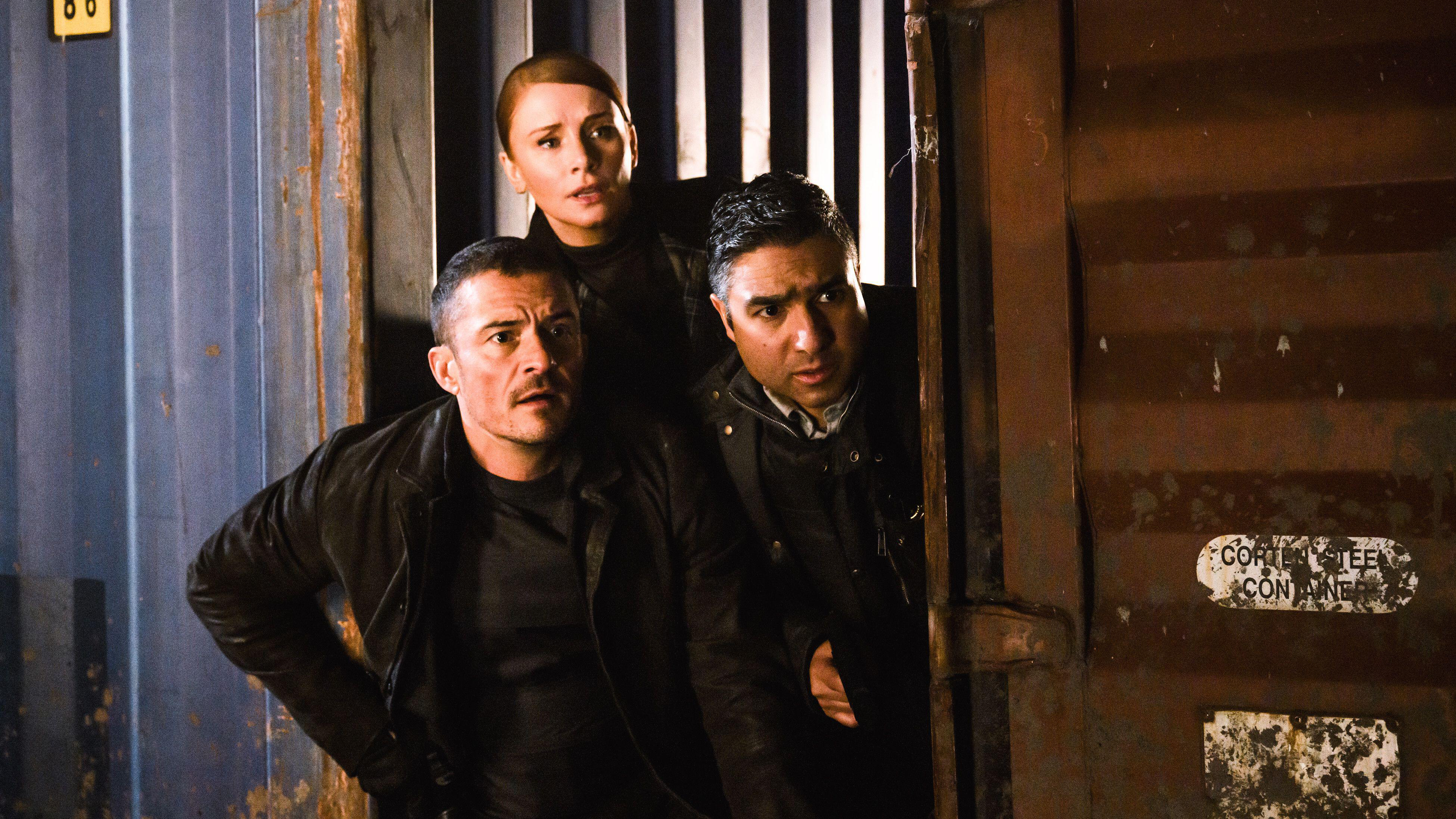 L-R: Marlon (Orlando Bloom), Kat (Bryce Dallas Howard) and Hugh (Nick Mohammed) lean around a shipping container together in a scene from Prime Video's "Deep Cover".
