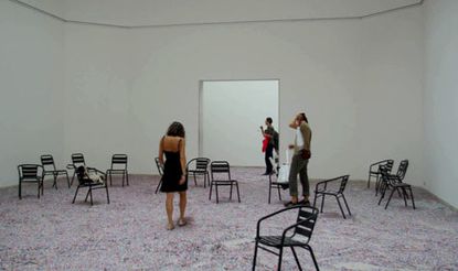 Room with white walls, multi coloured speckled floor and scattered black chairs, square opening to the room and three people walking towards the opening