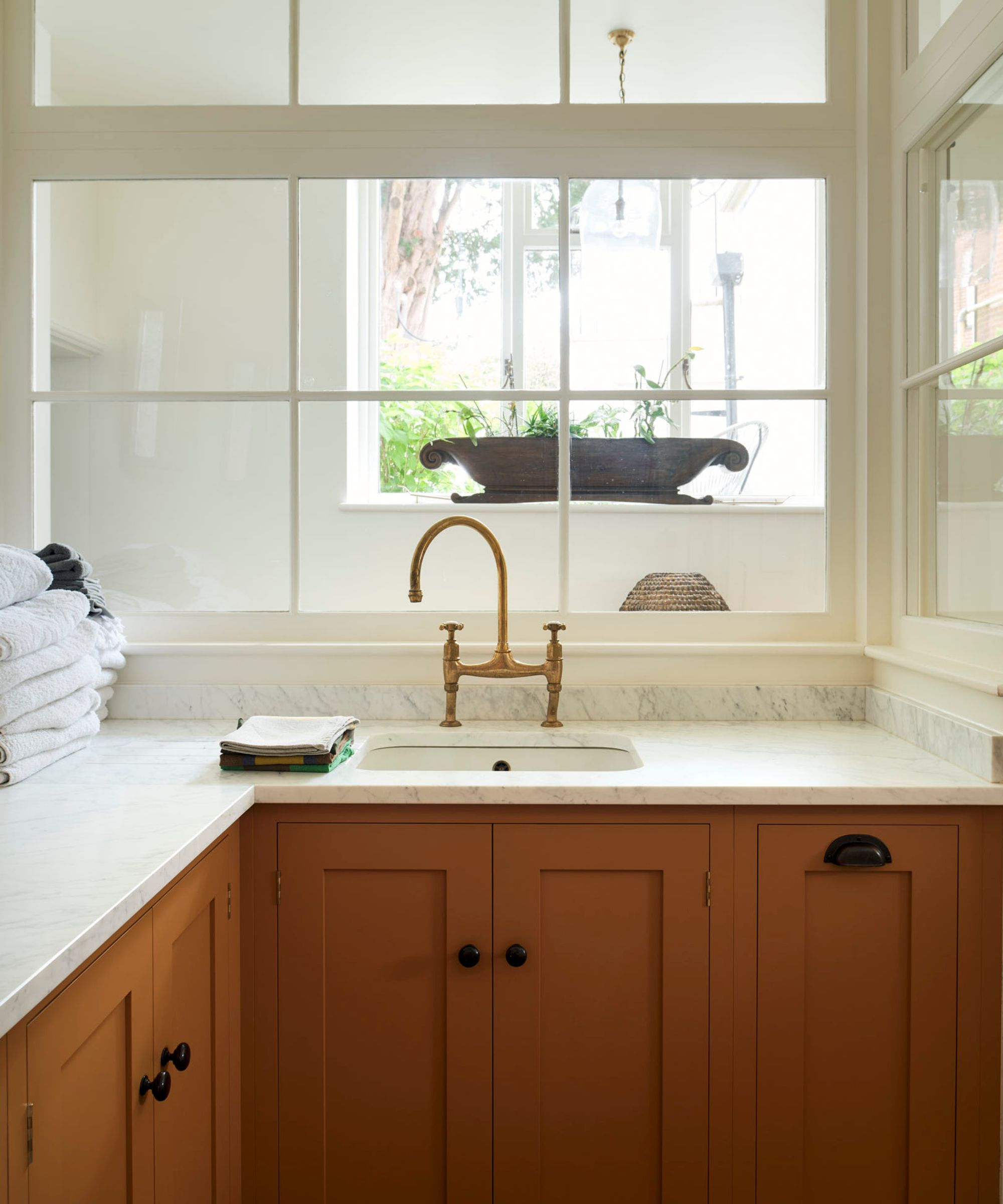 A compact utility room with burnt orange shaker-style cabinets, a marble worktop, brass mixer tap, and internal glass partition overlooking a bright window.