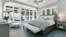 A bed with white and gray linens in a room with large windows looking out onto the water