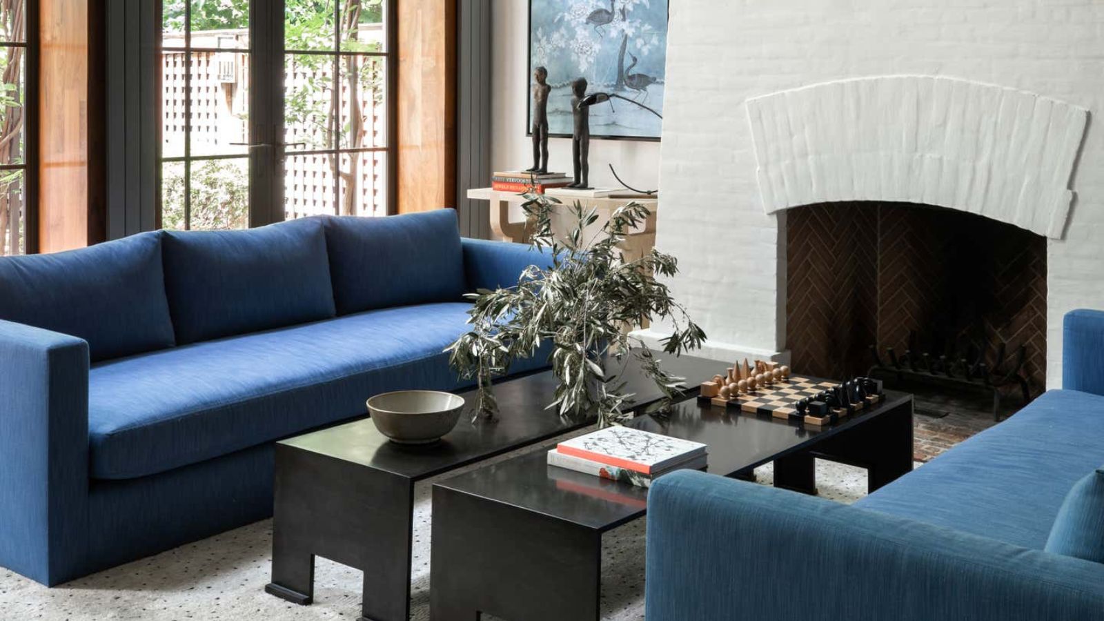 A living room with a large white-brick fireplace, two blue sofas, and a modern black metal coffee table between them. A plant, two books, a bowl, and a chessboard on the table. 