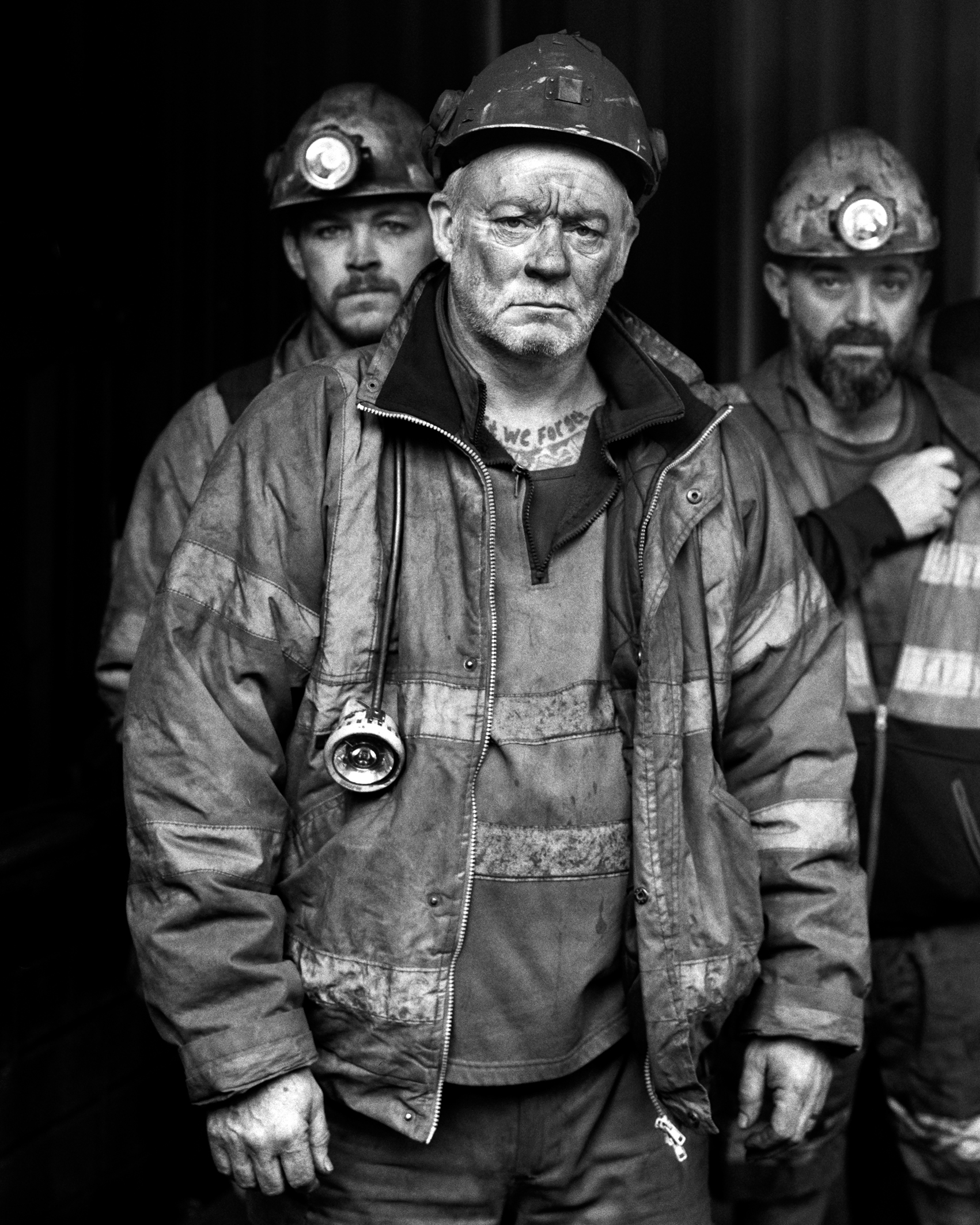 Black-and-white photo of three rugged coal miners wearing helmets and dirty work clothes, with serious expressions