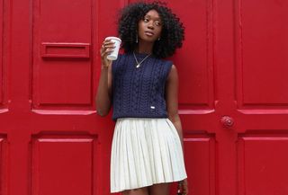 A woman wearing a white pleated miniskirt with a blue sweater vest at the start of a roundup of the best pleated miniskirts.