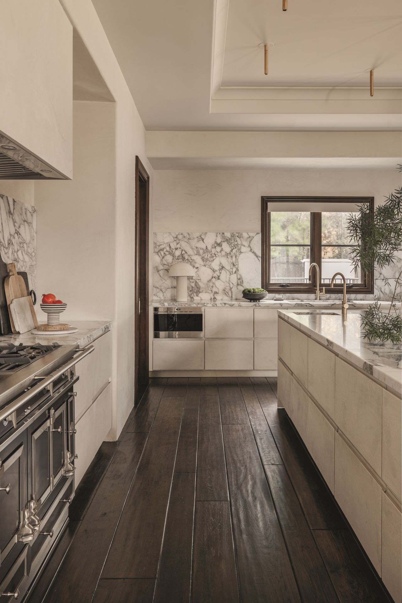 modern kitchen with beige walls, marble backsplash, dark timber floorboards, a table lamp on counter, brass taps, and cutting boards leaning against the backsplash