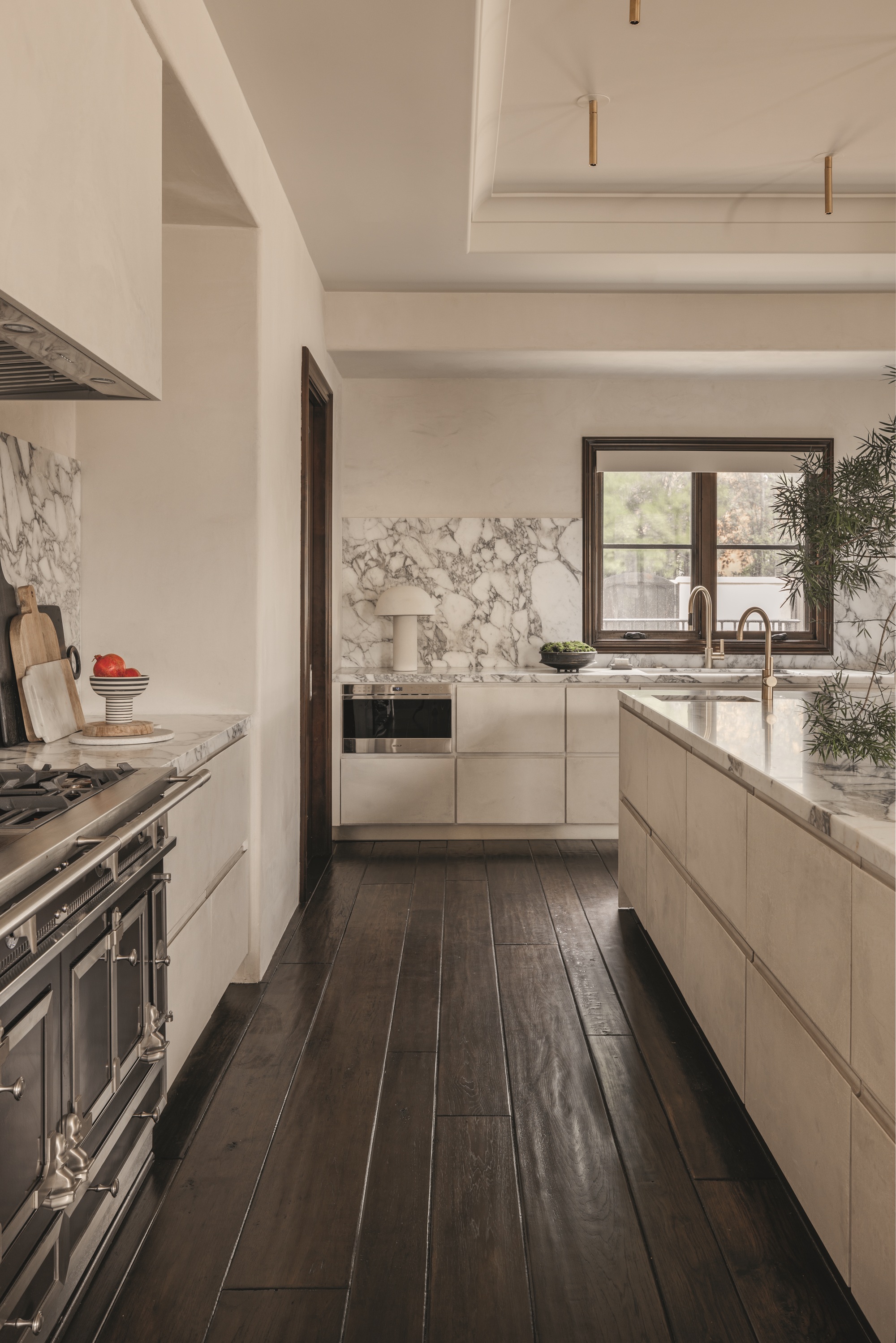 modern kitchen with beige walls, marble backsplash, dark timber floorboards, a table lamp on counter, brass taps, and cutting boards leaning against the backsplash