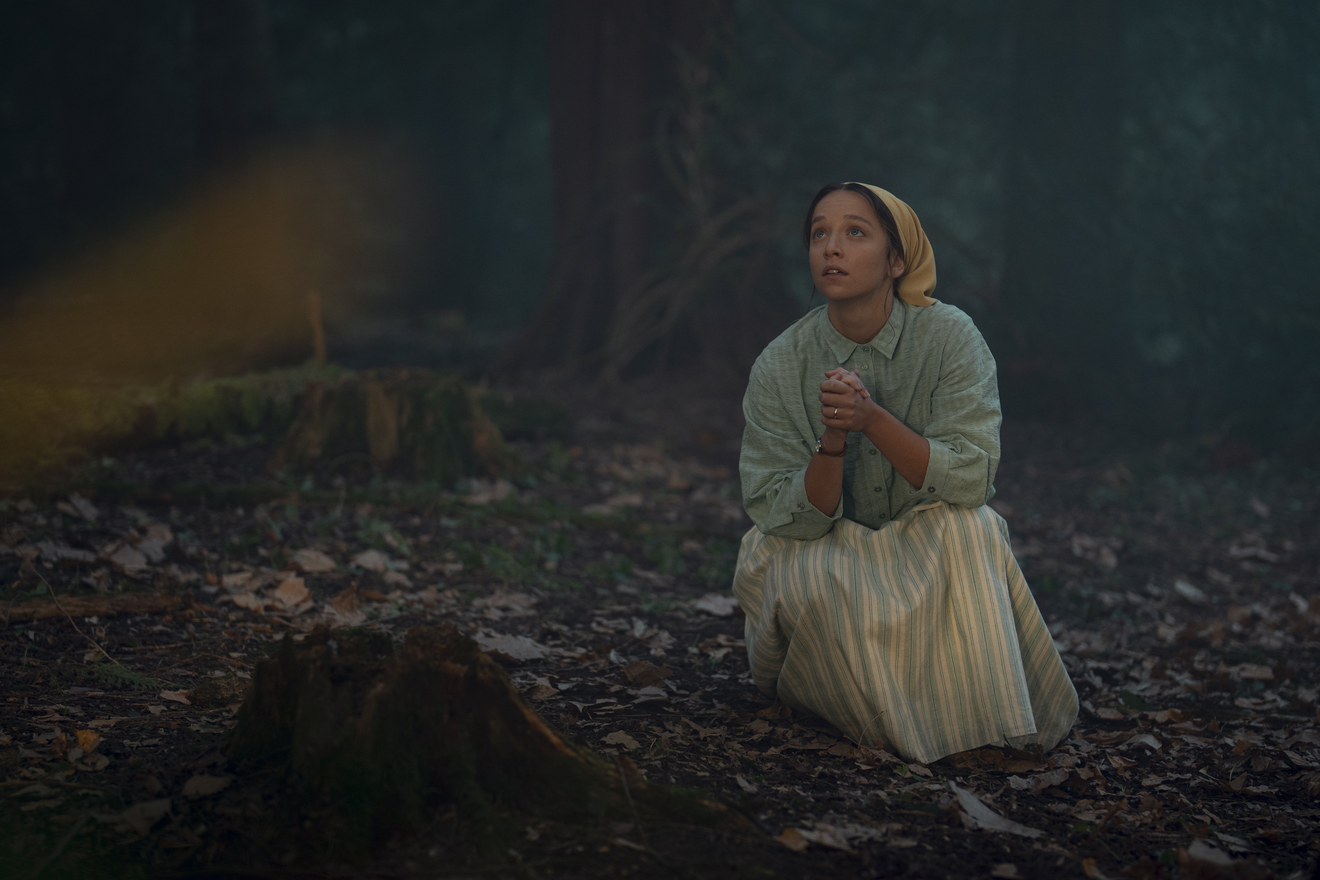 rosie crouches down to pray in a dark wood in a still from netflix's unchosen