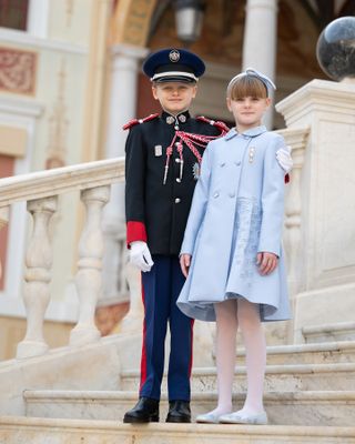 Prince Jacques wearing a military uniform and Princess Gabriella in a blue coat dress standing on the stairs of the royal palace in Monaco