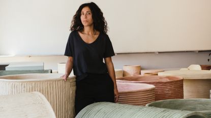 A young woman wearing a black loose-fitting T-shirt and trousers stands among her cement, pastel-shaded planters in a studio.