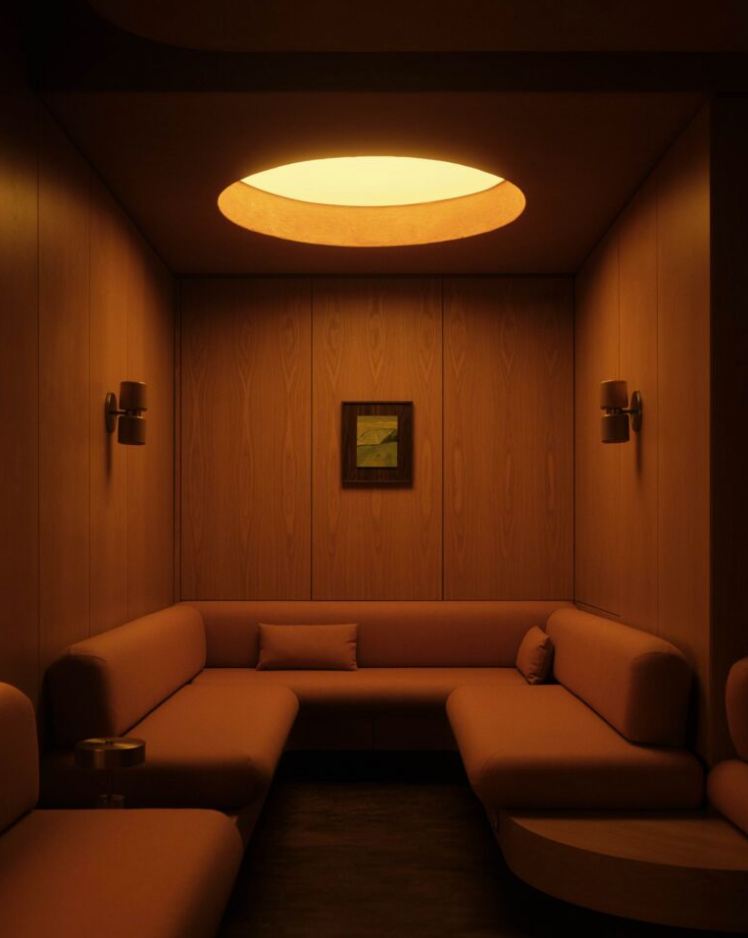 A cocooning restaurant booth with an U-shaped banquette in terracotta leather, wood-panelled walls, sconces, and one central artwork is overlooked by a golden-y ceiling porthole window.