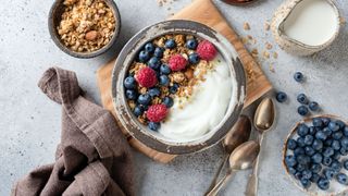 Bowl of yogurt and granola surrounded by various berries, nuts and healthy breakfast snacks