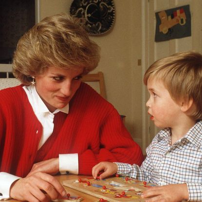 Princess Diana wearing a red cardigan over a white button up shirt sitting at a table playing a puzzle with a toddler Prince William