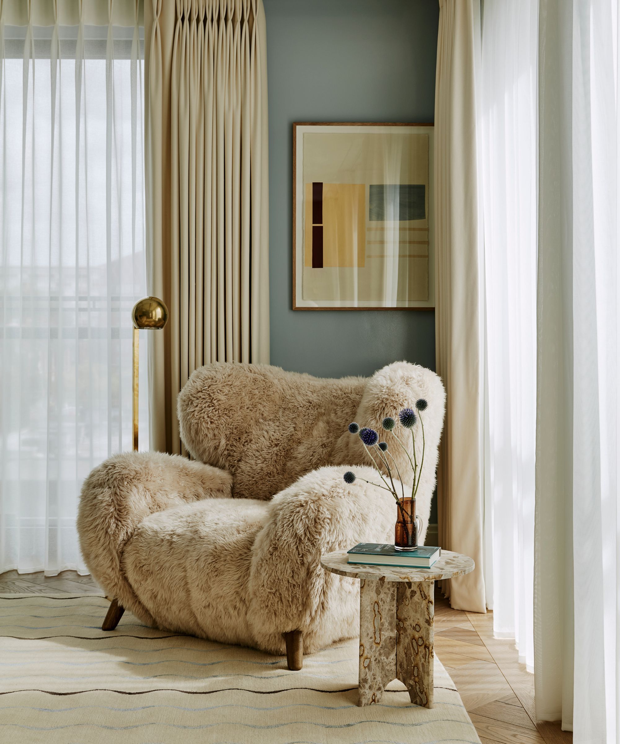 soft blue cozy modern bedroom with large windows, a neutral rug with wavy stripes, a fluffy sheepskin armchair styled with a mini marble table with a vase and a floor lamp